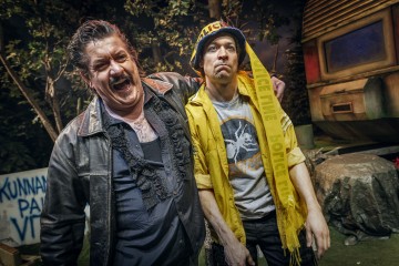 Kaksi miestä lavalla: toinen nauraa ja pitää toista käsivartensa toisen ympärillä, joka näyttää vakavalta ja jolla on poliisin hattu, varoitusnauha ja keltainen takki. Taustalla on viherkasveja ja kylttejä, jotka viittaavat teatteriesitykseen.