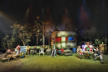 Ryhmä ihmisiä istuu ja seisoo metsäisessä ympäristössä olevan, säästä kärsivän perävaunun ympärillä, ja rekvisiitta, kuten tuolit, kylmälaukut ja pöytä, on valaistu värikkäillä näyttämövaloilla, jotka luovat vilkkaan ulkoilmakuvan.