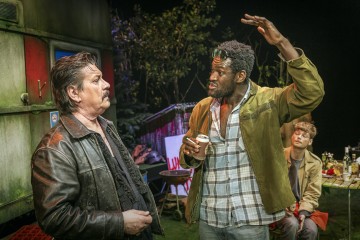 Kaksi miestä seisoo keskustelemassa; toinen elehtii vilkkaasti kohotetulla kädellä ja pitää tölkkiä kädessään. Molemmilla on takit. Kolmas mies istuu taustalla pullojen ja grillin keskellä. Ympäristö näyttää olevan ulkona, asuntovaunun lähellä.