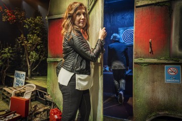 Mustaan takkiin pukeutunut nainen seisoo maalaismaisen, vihreän asuntovaunun ovella ja katsoo taaksepäin. Sisällä henkilö on tikkataulun edessä. Kohtaus on värikäs, taustalla on tupakointikieltokyltti ja puita.