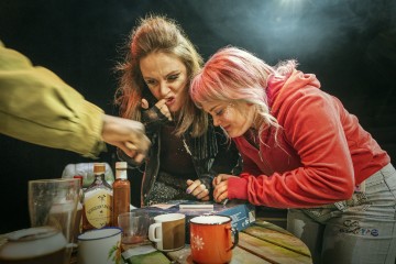 Kaksi naista nojaa pöydän yli, toinen nuolee sormeaan ja toinen hymyilee. Pöydällä on mukeja, pullo, laseja ja lautapeli. Kolmannen henkilön käsi osoittaa kädellä lautapeliä kohti. Kohtaus vaikuttaa leikkisältä ja vilkkaalta.