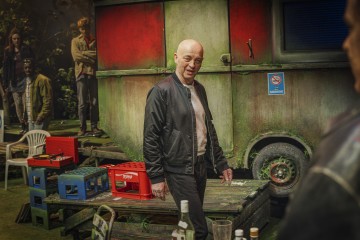 Mustaan takkiin pukeutunut kalju mies seisoo punavihreän ja sään värittämän asuntovaunun ulkopuolella, jota ympäröivät laatikot, pullot ja muovituolit. Taustalla on useita muita ihmisiä, jotka ovat osittain varjojen ja puiden peitossa.