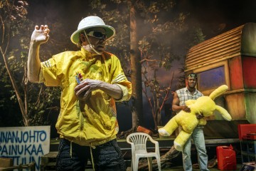 Keltaiseen takkiin, valkoiseen hattuun ja aurinkolaseihin pukeutunut henkilö pitelee nallea ja elehtii dramaattisesti, kun taas toinen aurinkolasipäinen henkilö seisoo takana pitelemässä suurta keltaista pehmolelua. Taustalla on puita ja kyltti.