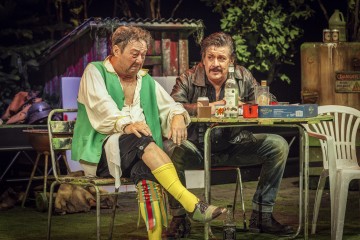 Kaksi miestä istuu sotkuisen ulkopöydän ääressä. Toisella on vihreä liivi, valkoinen paita ja keltaiset sukat, ja hän katsoo alakuloisesti. Toinen, tummaan takkiin pukeutunut, kumartuu puhuessaan. Pöydällä on erilaisia pulloja ja esineitä; taustalla on kasveja.