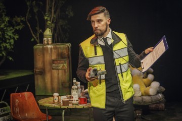 Keltaiseen heijastinliiviin pukeutunut mies pitää kameraa ja leikepöytää kädessään ja seisoo pullot, kupit ja tölkit täynnä olevan pöydän ääressä. Hänen takanaan on puita, vanha jääkaappi ja nalle sohvalla, mikä viittaa tutkinnan tapahtumapaikkaan.