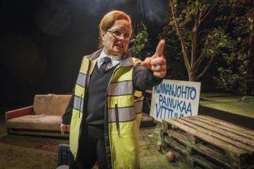 Silmälasit, heijastinliivit ja solmio päällään henkilö osoittaa painokkaasti lavalla, jossa on ulkotilojen rekvisiittaa, kuten sohva, puisia kuormalavoja ja suomenkielinen kyltti. Taustalla on puita ja lehtiä.