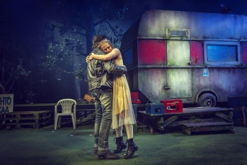 Nahkatakkinen mies ja vaaleaan mekkoon pukeutunut nainen syleilevät toisiaan tiukasti nurmikentällä, jonka taustalla on dramaattisessa valaistuksessa ränsistynyt asuntovaunu, muovituoleja ja laatikoita.