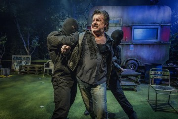 Kaksi mustaan pukeutunutta naamioitunutta hahmoa pidättelee mustapaitaista miestä ulkona olevan, säässä ränsistyneen asuntovaunun edessä, ja heidän ympärillään näkyy metsämaisemia ja näyttämörekvisiittaa.