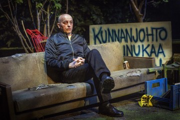 Mustaan takkiin pukeutunut kalju mies istuu yöllä kuluneella ulkosohvalla. Hänen takanaan on ostoskärry, laatikoita ja kyltti, jossa on sininen suomenkielinen teksti KUNNANJOHTO INUKAA. Puut ja hämärä valaistus luovat synkkää tunnelmaa.