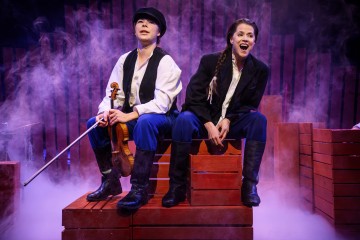 Kaksi aikakauden pukuihin pukeutunutta näyttelijää istuu punaisilla puulaatikoilla näyttämön sumun keskellä. Toisella on kädessään viulu ja päässään lippalakki, kun taas toinen, letitettynä, näyttää innostuneelta. Purppuravalaistu tausta viittaa dramaattiseen näyttämökohtaukseen.