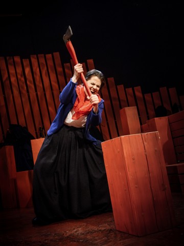 Dramaattisessa näyttämövalaistuksessa oleva henkilö nostaa kirveen päänsä yläpuolelle valmistautuessaan pilkkomaan punaista puupölkkyä intensiivinen ilme kasvoillaan. Taustalla on punaisia, abstrakteja puulevyjä.