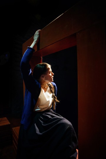 Siniseen villapaitaan ja tummaan hameeseen pukeutunut nainen istuu puurakenteella ja katsoo sivulle vakavalla ilmeellä, dramaattisesti valaistuna tummaa taustaa vasten.