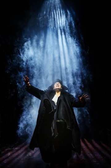 Henkilö seisoo dramaattisessa valaistuksessa kädet koholla ja suu auki, pyörteisen savun ympäröimänä ja yläpuolelta tulevan voimakkaan valonsäteen valaisemana, mikä luo teatraalisen ja intensiivisen tunnelman.