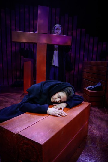 Mustiin pukeutunut nainen makaa pää lepää suurella punaisella puupalikalla silmät kiinni, kun taas toinen nainen seisoo suuren punaisen ristin takana ja katsoo synkkä ilme kasvoillaan hämärässä teatteriympäristössä.