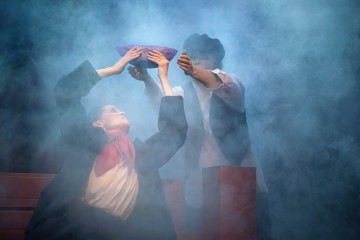 Kaksi näyttelijää esiintyy lavalla paksun sinisen sumun keskellä; toinen nostaa violetin kulhon päänsä yläpuolelle, kun taas toinen kurottautuu sitä kohti. Molemmilla on tummat, dramaattiset puvut, ja kohtaus vaikuttaa intensiiviseltä ja teatraaliselta.