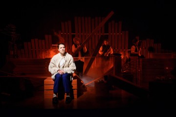 Valkoiseen villapaitaan pukeutunut henkilö istuu penkillä hämärästi valaistun näyttämön edessä, ja taustalla esiintyy kolme ihmistä punaisen valaistuksen ja puisten lavasteiden keskellä, jotka luovat dramaattisen tunnelman.