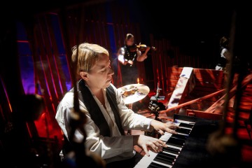 Muusikko soittaa lavalla näppäimistöä keskittyneesti, dramaattisessa punaisessa ja sinisessä valaistuksessa. Taustalla soittavat rumpali ja viulisti, jotka luovat tunnelmallisen konserttiympäristön.