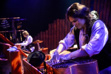 Pitkätukkainen muusikko soittaa jousisoitinta näyttämöllä dramaattisessa valaistuksessa, kun toinen muusikko esiintyy taustalla punaisen näyttämörekvisiitan ja instrumenttien keskellä.