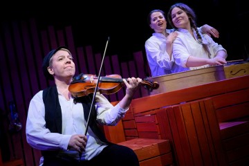 Muusikko soittaa viulua etualalla, kun kaksi valkoisiin paitoihin pukeutunutta naista laulaa yhdessä hänen takanaan istuen punaisella puisella lavasteella dramaattisessa näyttämövalaistuksessa.