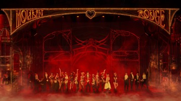 Suuri joukko esiintyjiä seisoo lavalla eloisan punaisen ja kultaisen valaistuksen alla, jota kehystää koristeellinen Moulin Rouge -kyltti, ja taidokas lavastus tuo mieleen vilkkaan kabareetunnelman.