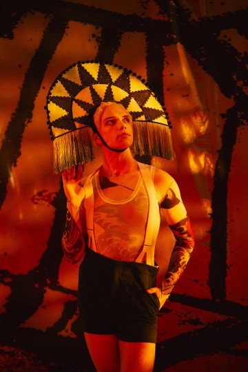 Henkilö, jolla on dramaattinen, mustavalkoinen hapsullinen päähine, läpikuultava hihaton toppi, mustat shortsit, joissa on henkselit, ja käsivarretatuoinnit, poseeraa itsevarmasti oranssia abstraktia taustaa vasten.