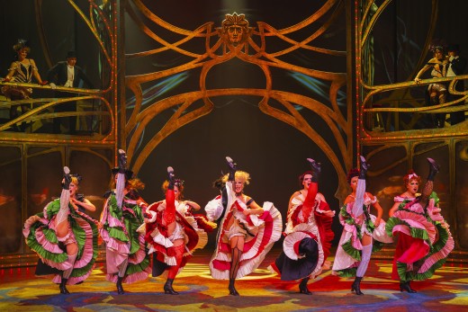 Seitsemän tanssijaa värikkäissä can-can-puvuissa potkivat jalkojaan korkealle yhdessä eloisalla näyttämöllä, jota kehystävät koristeelliset kultaiset kaaret ja dramaattinen valaistus, ja esiintyjät katselevat taustalla olevilta korotetuilta lavoilta.