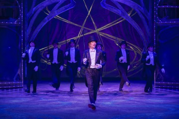 Joukko esiintyjiä smokkeihin, silinterihattuihin ja valkoisiin hanskoihin pukeutuneina tanssii muodostelmassa kirkkaasti valaistulla lavalla, jonka taustana on violetti ja kultainen tausta ja taidokkaat kaarikuviot. Päätanssija on etualalla ja laulaa tai puhuu.