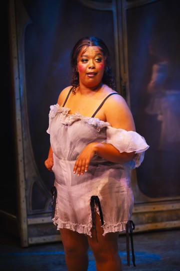 Henkilö, jolla on näyttämömeikki ja läpikuultava, olkapäätön valkoinen puku, seisoo dramaattisessa valaistuksessa tumman, teatterimaisen taustan edessä. Hänen ilmeensä vaikuttaa mietteliäältä ja esitykseen sitoutuneelta.