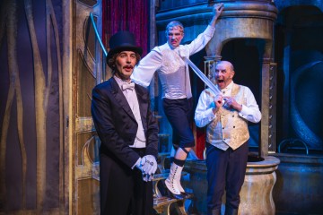 Lavalla esiintyy kolme teatteripukuista miestä; yksi mustaan smokkiin ja silinterihattuun pukeutuneista seisoo edessä, kun taas kaksi muuta valkoisiin ja kultaisiin liiveihin pukeutunutta miestä on vilkkaasti vuorovaikutuksessa punaisilla verhoilla varustetun metallisen lavastuksen lähellä.