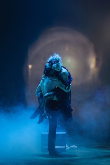 Paidaton henkilö kantaa toista henkilöä dramaattisessa poseerauksessa sinisen näyttämövalaistuksen ja sumun ympäröimänä. Taustalla on hohtava kaari, joka viittaa teatteri- tai tanssiesitykseen.