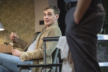 Ruskehtavaan takkiin ja farkkuihin pukeutunut mies istuu rennosti, viinilasi vieressä pöydällä, pitää silmälasejaan ja katsoo toista lähellä seisovaa henkilöä. Taustalla on vihreäkuvioinen seinä.