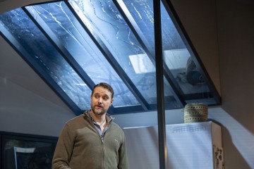 Lyhythiuksinen ja parrakas mies seisoo sisätiloissa suuren ikkunakattorakenteen lähellä, jonka lasissa on sadepisaroita. Hänellä on harmaa vetoketjullinen villapaita ja hän näyttää mietteliäältä. Taustalla näkyy jääkaappi ja punottuja koreja.