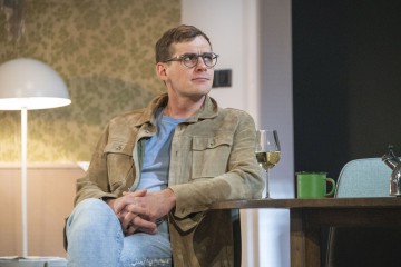 Silmälasipäinen, ruskehtava takki ja farkut jalka ristissä istuu pöydän ääressä, jossa on lasi valkoviiniä, vihreä muki ja teekannu. Taustalla on kuviollinen tapetti ja moderni lattiavalaisin.