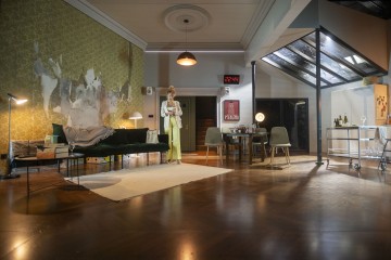 Nainen seisoo tyylikkäässä, modernissa olohuoneessa, jossa on vihreä sohva, vintage-tapetti, ruokapöytä, seinäkello, jossa lukema on 22:24, ja lämmin valaistus, joka luo viihtyisän tunnelman.