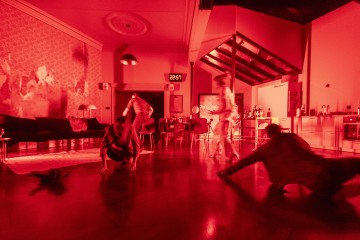 Hämärästi valaistussa, punaisessa valossa kylpevässä huoneessa näkyy neljä sumeaa hahmoa, jotka liikkuvat puulattialla, ja taustalla on huonekaluja, keittiö ja digitaalikello, jossa lukee 22:51.