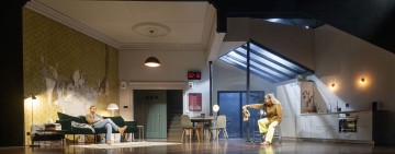 Moderni olohuone ja keittiö, jossa yksi henkilö rentoutuu vihreällä sohvalla ja toinen istuu ruokapöydän ääressä ja elehtii esine kädessään. Seinällä oleva digitaalikello näyttää kello 12:16.