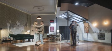 Kaksi pariskuntaa syleilee modernissa olohuoneessa ja keittiössä, joka on lavastettu näyttämölle. Huoneessa on vihreä sohva, ruokapöytä, suuri kello seinällä ja nykyaikainen sisustus lämpimässä, dramaattisessa valaistuksessa.