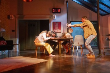 Henkilö seisoo kädet ojennettuina kohti kolmea pöydän päälle lyyhistynyttä ihmistä modernin olohuoneen dramaattisessa valaistuksessa; digitaalikellossa lukee 01:18 ja 