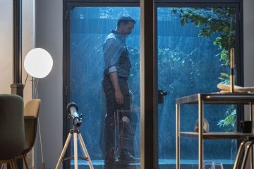 Mies seisoo sisätiloissa lasioven luona yöllä ja katsoo alaspäin vakavalla ilmeellä. Ulkona sataa rankasti. Modernissa huoneessa näkyy kaukoputki, pyöreä lamppu ja ruokapöytä.