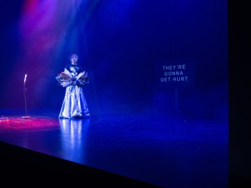 Dramaattiseen pukuun pukeutunut esiintyjä seisoo hämärästi valaistulla näyttämöllä kukkakimppu kädessään, ja yläpuolella on värikkäitä valoja. Mikrofoni on lähellä, ja taustalla olevalla näytöllä näkyy teksti THEYRE GONNA GET HURT.