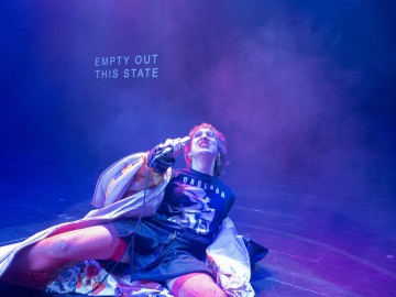 Esiintyjä istuu lattialla mikrofonin kanssa kirkkaassa näyttämövalaistuksessa ja savussa dramaattisessa asussa ja punaisissa verkkosukissa. Taustalle heijastettu teksti kuuluu: EMPTY OUT THIS STATE.