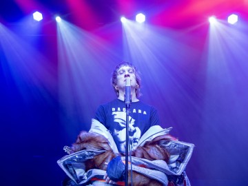 Laulaja seisoo lavalla dramaattisessa valaistuksessa, yllään suuri, värikäs takki ja musta graafinen t-paita, ja hänen takanaan loistavat kirkkaan siniset ja vaaleanpunaiset lavavalot.