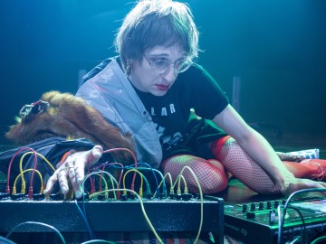 Lyhythiuksinen, silmälasipäinen ja punaisissa verkkosukissa oleva henkilö säätää sähköisten musiikkilaitteiden kaapeleita ja säätimiä sinisen lavavalaistuksen alla. Hänen olkapäänsä yli on ripustettu turkisasuste.