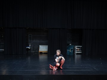 Mustaan paitaan ja punaisiin sukkahousuihin pukeutunut henkilö kyyristyy keskellä pimeää, tyhjää teatterilavaa, jossa on mustat verhot. Rikkinäistä punaista rekvisiittaa tai puvun osia on hajallaan heidän lähellään. Taustalla on minimaalisesti huonekaluja.