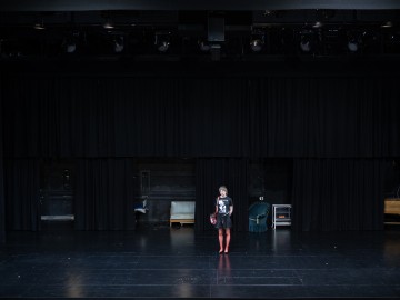 Yksinäinen henkilö seisoo keskellä näyttämöä mustien verhojen edessä hämärästi valaistussa teatterissa, yllään musta asu ja punaiset kengät. Taustalla näkyy näyttämötarvikkeita, kuten penkki ja katettuja esineitä.