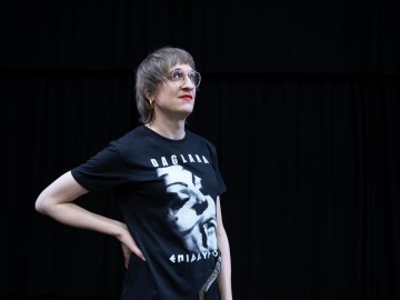 Lyhythiuksinen, silmälasipäinen ja punaisella huulipunalla varustettu henkilö seisoo toinen käsi lantiollaan, päällään musta graafinen t-paita, mustan verhotaustan edessä.