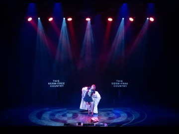 Esiintyjä seisoo näyttämön keskellä värikkäiden valonheittimien alla valkoiseen lakanaan käärittynä. Kahdella näytöllä molemmin puolin näkyy teksti THIS GERM-FREE COUNTRY. Lavalla on hämärä valaistus ja dramaattinen tunnelma.