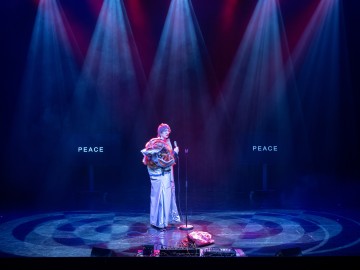 Hopeanvärisiin kiiltäviin vaatteisiin pukeutunut esiintyjä seisoo lavalla sinisten ja punaisten valonheittimien alla, kädessään värikkäitä rekvisiittaa, ja hänen takanaan on kaksi näyttöä, joilla lukee sana PEACE.