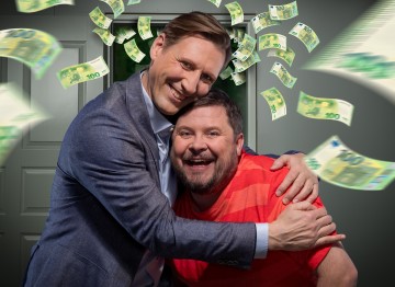 Kaksi hymyilevää miestä halaa avoimen oven edessä, toisella on puku ja toisella punainen raitapaita. Heidän ympärilleen näyttää satelevan kymmeniä 100 euron seteleitä.