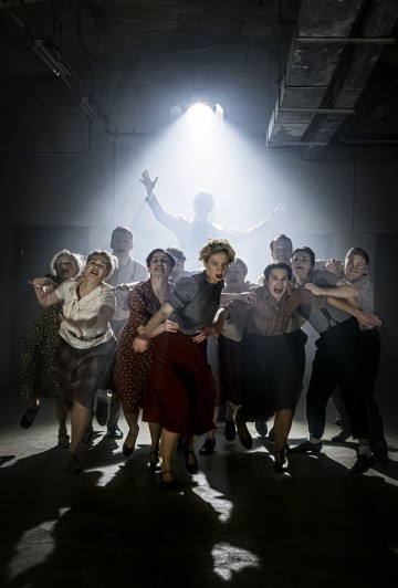 Ryhmä vintage-vaatteisiin pukeutuneita ihmisiä tanssii tarmokkaasti eteenpäin dramaattisessa näyttämövalaistuksessa, kun yksi henkilö valaistaan taustalla, kädet koholla ja savu tai sumu luo teatteritunnelmaa.